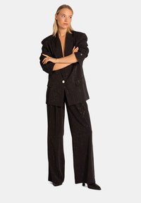 Zwart gestreept pak met een op maat gemaakt blazer met een enkele zilveren knop en wijde broekspijpen; de stof heeft een gestructureerde afwerking met subtiele glans.