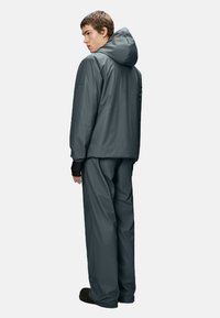 Grijze waterdichte jas met capuchon, voorzien van een losse pasvorm en elastische manchetten, gecombineerd met bijpassende grijze broek en zwarte schoenen.