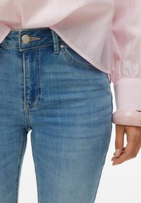 Išblukę mėlyni džinsai iš džinsinio audinio su aukšta liemeniu, kontrastinėmis siūlėmis ir priekyje užsegami mygtukais. Derinami su šviesiai rožine tekstūruota palaidine.