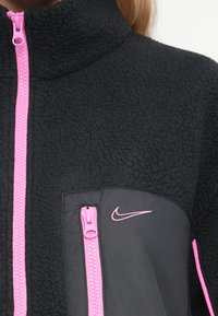 Crna jakna od flisa s visokim ovratnikom, koja ima ružičaste zatvarače, zatvoreni džep na prsima i mali Nike logotip na džepu.