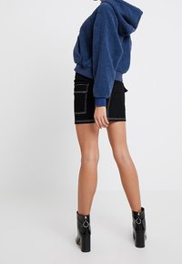 Sudadera de forro polar azul con capucha, combinada con una minifalda de mezclilla negra con costuras blancas y bolsillos, complementada con botas negras brillantes hasta el tobillo.