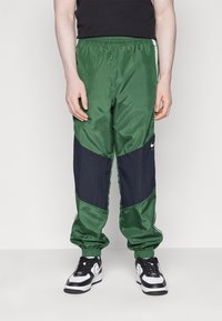 Groene en marineblauwe sportbroek met een glanzende textuur, elastische tailleband en contrasterend panneldesign. Zwarte Nike-logo zichtbaar aan de zijkant.