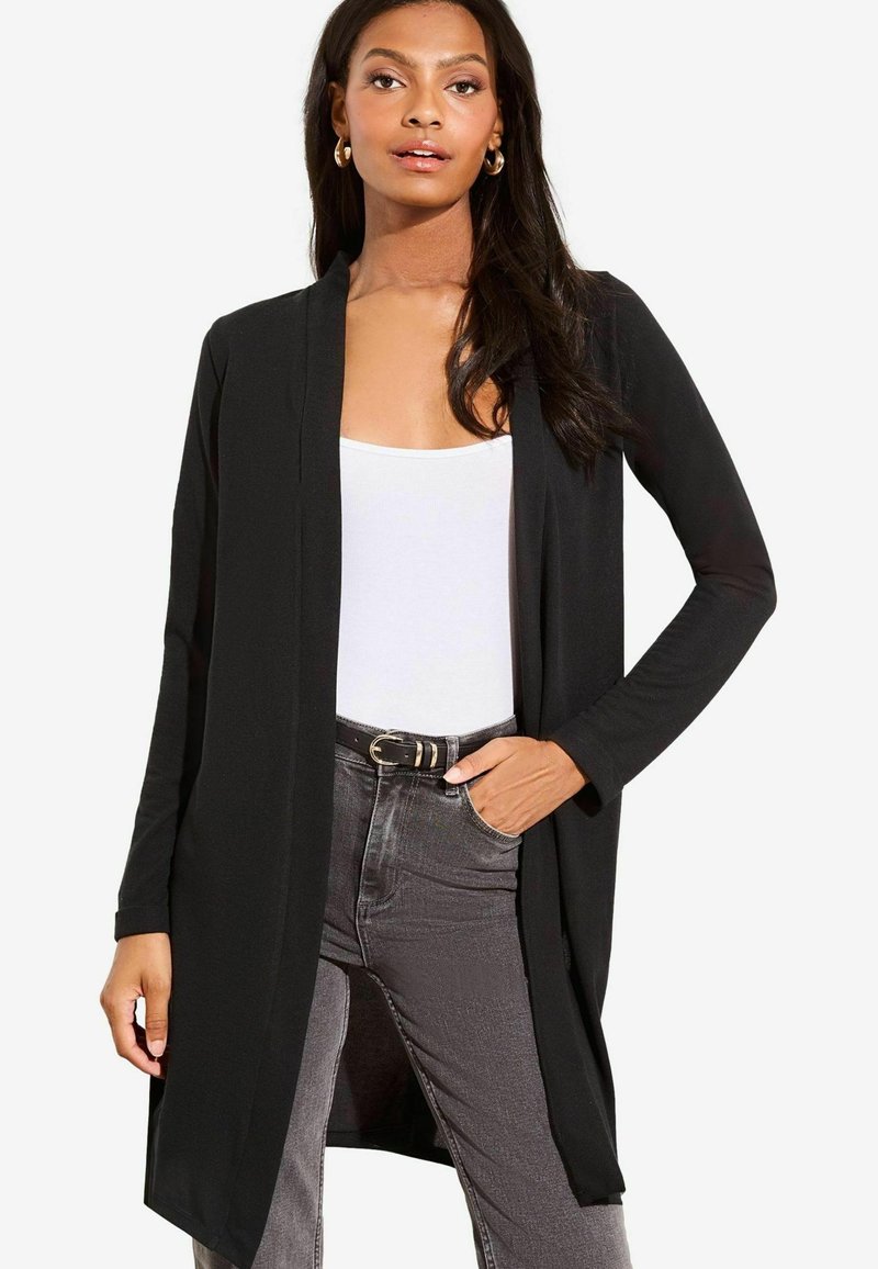Gilet ouvert noir à manches longues, en tissu doux, avec une coupe décontractée et des fentes sur les côtés, porté sur un haut blanc et un jean gris.