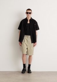 Camisa negra de manga corta sobre una camiseta negra sin mangas, combinada con pantalones cortos beige. Los accesorios incluyen gafas de sol negras y zapatos con calcetines negros.