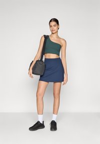 Roheline ühekätine crop top, sinine seelik ja mustad tossud. Mudel kannab musta kotti. Lihtne disain, kehakohane siluett ja sile kangas.