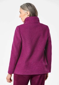 Paarse wolmixjas met een hoge kraag, een op maat gemaakt ontwerp en zichtbare naaddetails; gedragen over bijpassende paarse broeken.