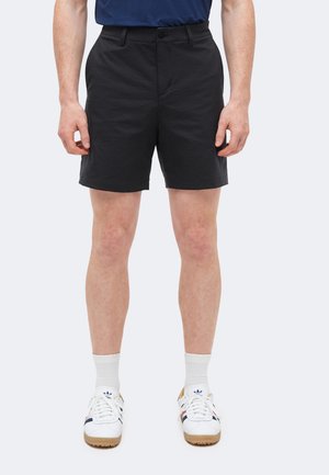 Mężczyzna w czarnych krótkich spodenkach do kolan, białych skarpetach i białych butach sportowych Adidas z granatowymi i czerwonymi paskami.