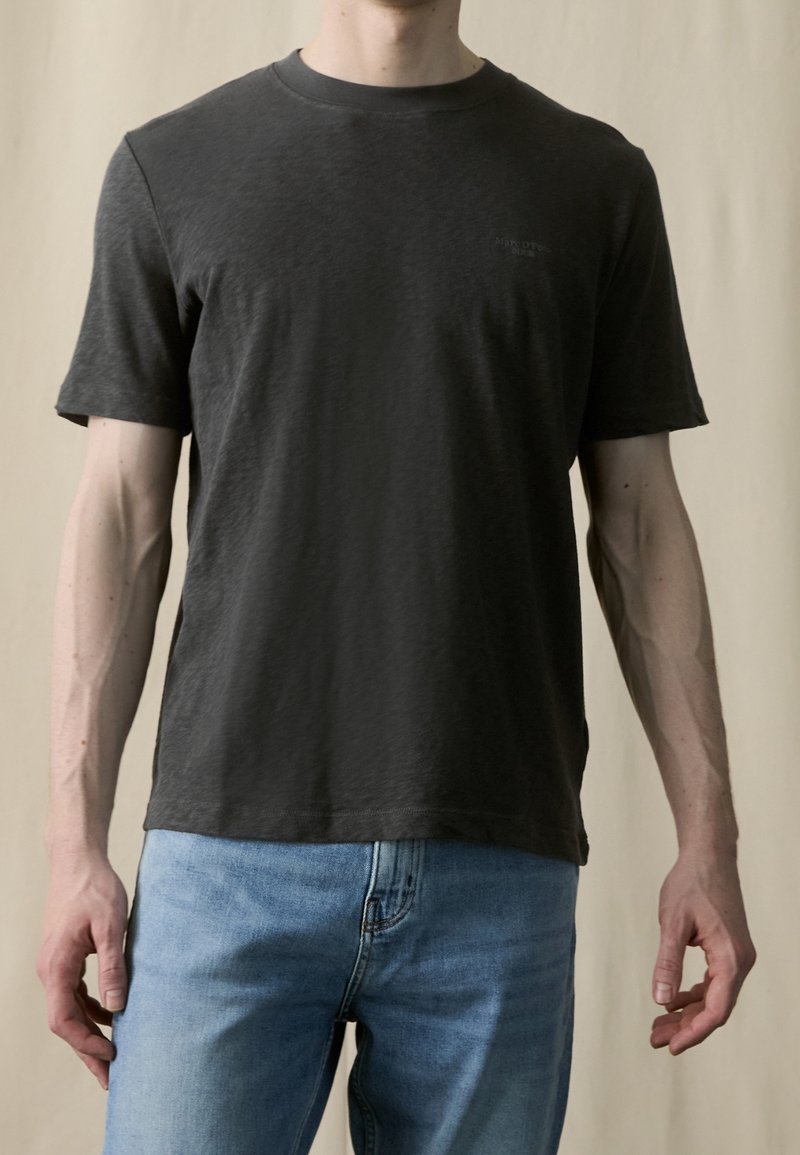 Camiseta de manga corta de algodón en color gris oscuro con cuello redondo. Presenta un pequeño logo en el pecho izquierdo y un corte holgado. Combinada con vaqueros azules.