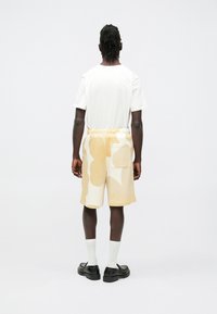 Pantalones cortos con un patrón abstracto en crema y beige, cintura elástica, bolsillo lateral, combinados con una camiseta blanca y zapatos negros.