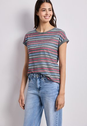 Kobieta uśmiecha się, ubrana w szary T-shirt z krótkim rękawem z różowymi i niebieskimi paskami oraz jasnoniebieskie dżinsy z wysokim stanem na jednolitym tle.