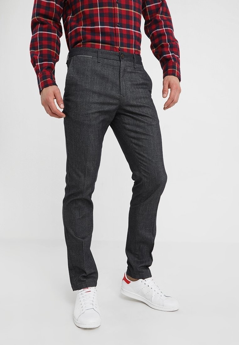 Pantalon gris foncé ajusté en tissu texturé, avec un devant plat, des poches latérales et une boucle de ceinture discrète. Porté avec des baskets blanches.