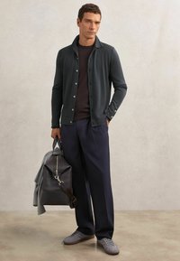 Dunkelgrüner Cardigan mit Knöpfen, getragen über einem dunklen T-Shirt, kombiniert mit marineblauer Hose und hellgrauen Sneakers; trägt eine graue Reisetasche.