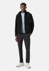 Schwarze Jacke mit hohem Kragen, hellblauer Pullover darunter, dunkle Hose und weiße Turnschuhe. Einfaches Design, glatte Textur und funktionale Details.