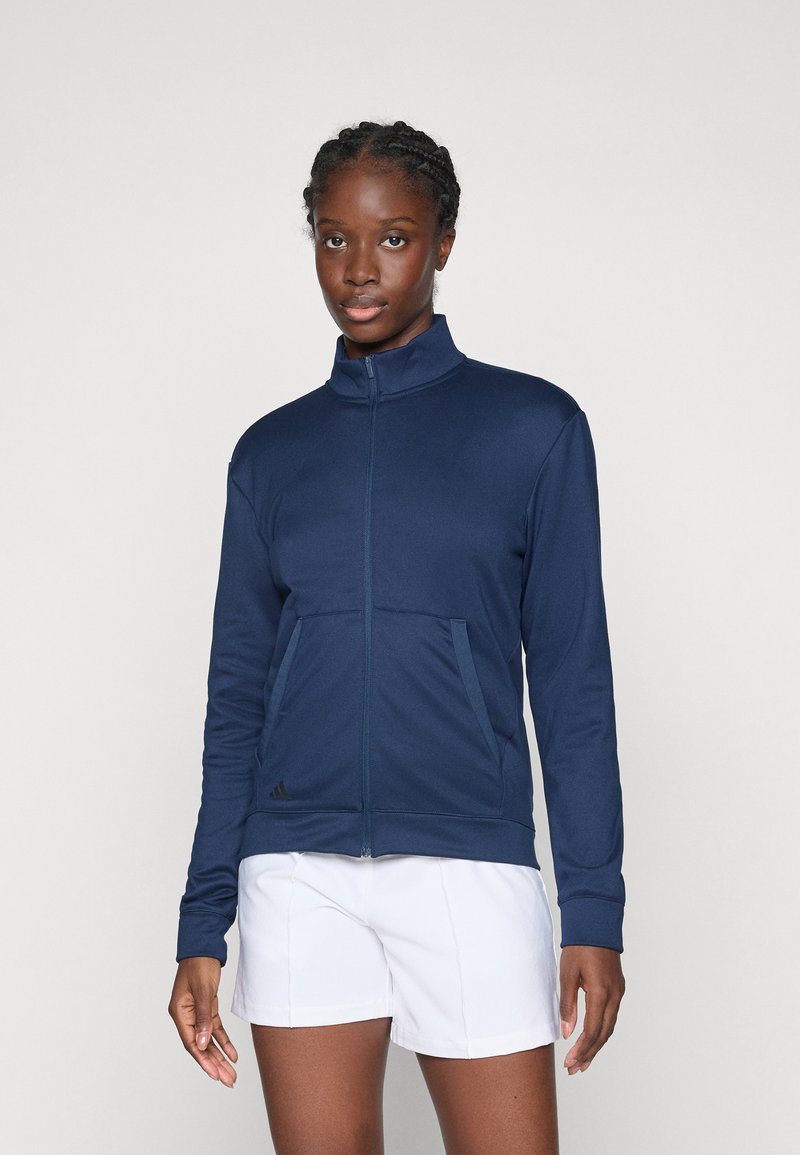 Woman with braided hair wears a navy zip-up jacket and white shorts, standing against a plain light gray background.