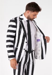 Čierno-biely pruhovaný oblek s lesklým povrchom. Sako má vzor s rozliatimi škvrnami a podšívka zobrazuje tému Beetlejuice.