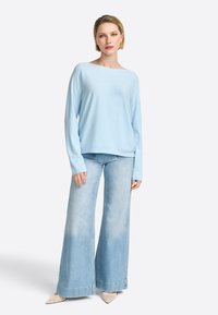 Hellblaue, langärmelige Strickjacke kombiniert mit weiten, ausgebleichten Jeans. Die Strickjacke hat eine entspannte Passform und einen Boot-Ausschnitt; die Jeans haben einen fransigen Saum.
