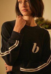 Zwarte sweatshirt met witte en gouden gestreepte mouwen, ronde halslijn en een geborduurd "b"-logo op de borst. Zachte stofstructuur.