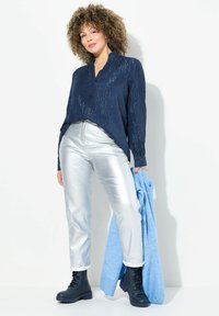 Camisa de manga larga azul marino con rayas verticales, pantalones metálicos plateados, botas negras con cordones y una chaqueta azul claro colgada del brazo.
