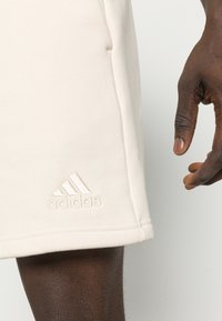 Beżowe szorty Adidas wykonane z miękkiego materiału, z wytłoczonym logo na krawędzi. Gładka faktura i luźny krój.