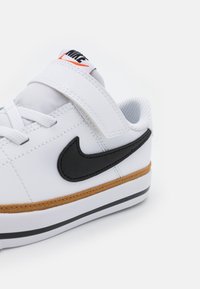 White sneaker with a black swoosh logo, textile and synthetic upper, padded strap, brown midsole, and black accents on the rubber outsole.