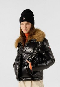 Schwarze Pufferjacke mit einem mit Fell gefütterten Kapuzen, glänzender Textur, Reißverschluss und orangem Akzent am Bündchen. Schwarze Strickmütze mit Logo-Patch.