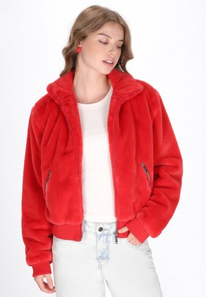 Bomber jacket in finta pelliccia rossa con colletto alto, tasche con zip e polsini a coste. Indossato sopra una camicia bianca e jeans azzurri chiari.