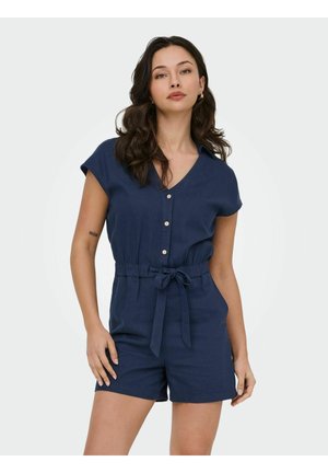 Femme aux longs cheveux foncés porte une combinaison courte bleu marine à manches courtes avec boutons, col en V et ceinture nouée à la taille, debout devant un fond uni.