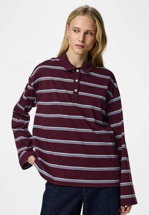 Jauna sieviete ar gariem blondiem matiem, tērpusies bordo krāsas garroku polo kreklā ar baltām horizontālām svītrām un tumši zilās džinsās, stāv ar roku kabatā.