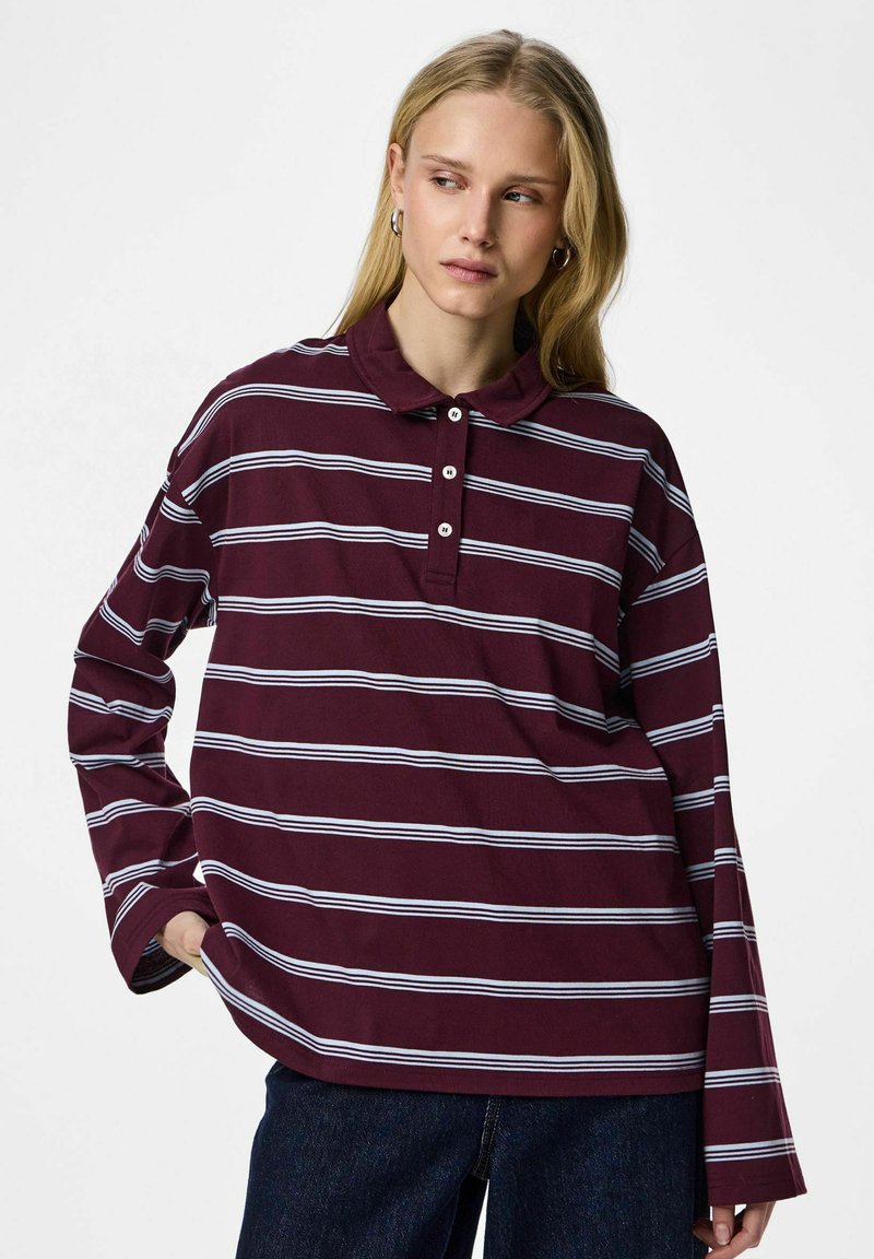 Ung kvinde med langt blondt hår iført en vinrød polotrøje med lange ærmer og hvide vandrette striber samt mørkeblå jeans, stående med hånden i lommen.