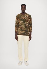 Gebreide trui in aardse tinten met een patronenlandschapdesign, gecombineerd met crèmekleurige broeken en grijze sneakers.