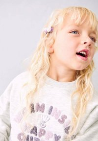 Bambino biondo con gli occhi azzurri che indossa un maglione chiaro con lettere in rilievo e una piccola clip per capelli color lavanda, che guarda in alto e a destra.