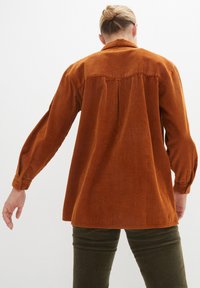 Chemise en velours côtelé couleur orange brûlé avec une coupe décontractée, dotée de manches longues et de détails en côtes verticales sur le tissu.