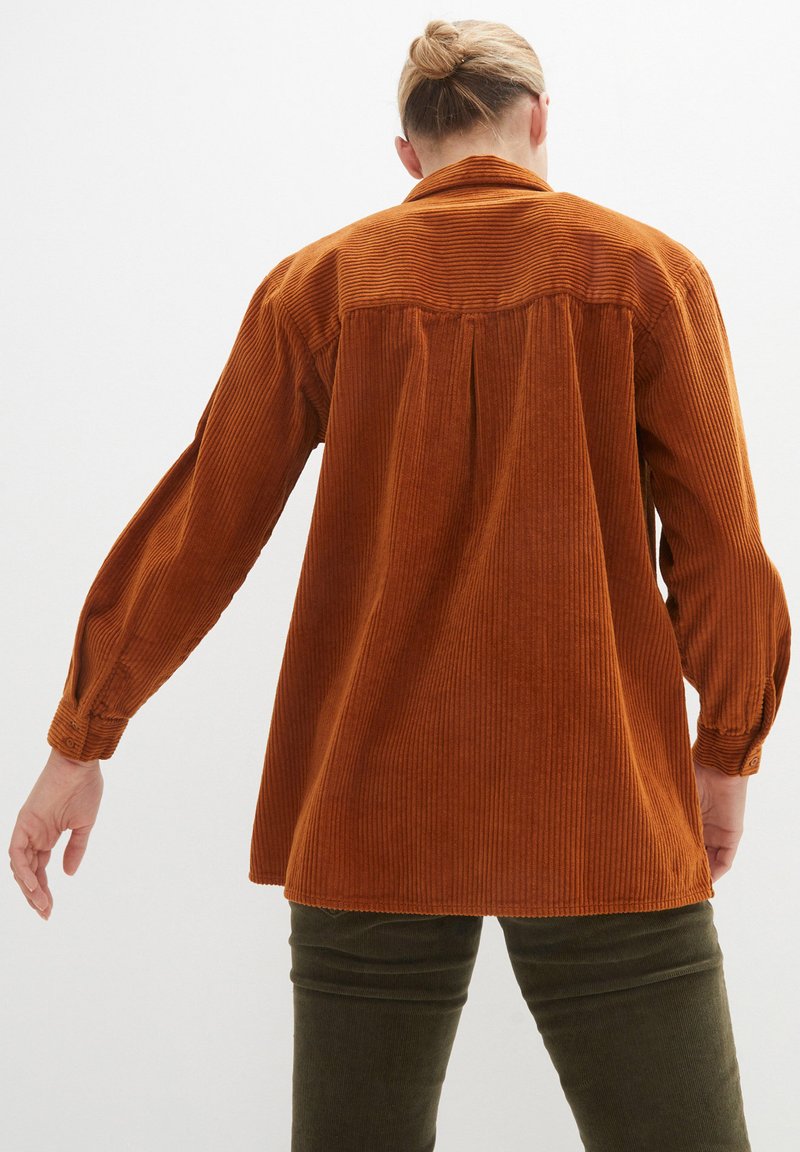 Chemise en velours côtelé couleur orange brûlé avec une coupe décontractée, dotée de manches longues et de détails en côtes verticales sur le tissu.