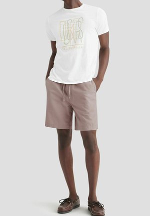 Man met een wit t-shirt met grafische print, lichtbruine short met trekkoord en donkerbruine bootschoenen, staand met handen in de zakken.