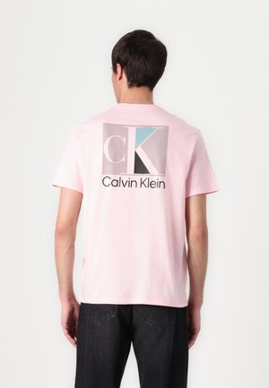 Ein Mann, der von hinten zu sehen ist, trägt ein hellrosa Calvin Klein T-Shirt mit einem großen, mehrfarbigen CK-Logo und Text auf dem Rücken, kombiniert mit einer schwarzen Hose.
