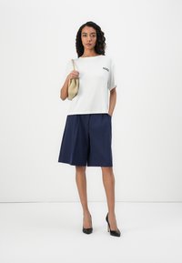 Vit kortärmad T-shirt med "HUGO"-logotyp, matchad med marinblå veckade shorts och svarta högklackade skor, hållande en beige handväska.