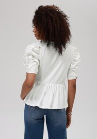 White short-sleeve top with puffed sleeves, gathered waist, and a flared peplum hem. Paired with blue denim jeans. Smooth fabric texture.