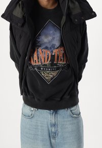 Persona che indossa una felpa nera con una grafica montana "Grand Teton Wyoming", un giubbotto senza maniche nero e jeans azzurri chiari.