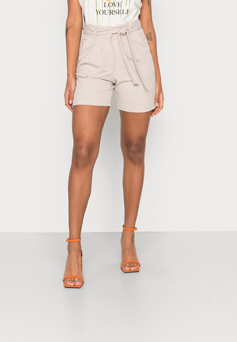 Femme portant un short beige taille haute ceinturé, un haut blanc avec l'inscription « Love Yourself » et des sandales à talons hauts orange, debout devant un fond uni.