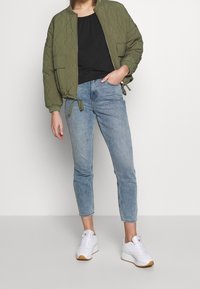 Kvinna i olivgrön vadderad bomberjacka, svart topp, ljusblå cropped jeans och vita sneakers mot en enfärgad bakgrund.