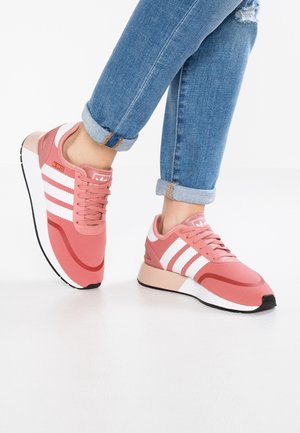 Person iført lyserøde Adidas-sneakers med hvide striber og opbukket blå jeans, stående på en hvid gulv mod en ensfarvet baggrund.