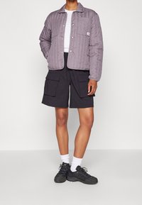 Ljuslila kviltad jacka med tryckknappar, matchad med svarta cargoshorts. Modellen bär svarta sportskor och vita strumpor.