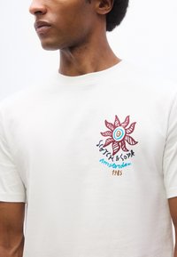 Άνδρας που φοράει λευκό T-shirt με κόκκινο γραφικό ήλιο και κείμενο «Scotch & Soda Amsterdam 1985» στο πάνω αριστερό μέρος του στήθους.