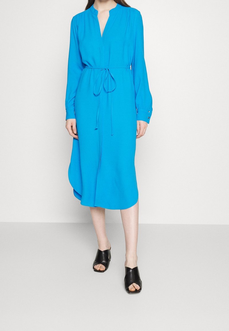 Vrouw draagt een felblauwe midi-jurk met lange mouwen, een tailleband en zwarte open-toe sandalen met hak, staand tegen een effen achtergrond.