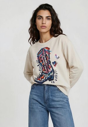 Woman with dark wavy hair wearing a beige sweatshirt with a colorful cowboy boot and hearts print, paired with blue high-waisted jeans.