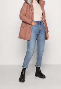 Brązowa ocieplana parka z futrzanym kapturem, zestawiona z jasnobłękitnymi spodniami o wysokim stanie i czarnymi botkami na kostkę z grubą podeszwą.