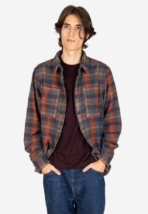 Junger Mann mit mittellangen Haaren, der ein rot-blau-braunes kariertes Hemd über einem dunklen T-Shirt und blauen Jeans trägt, die Hände in den Taschen.