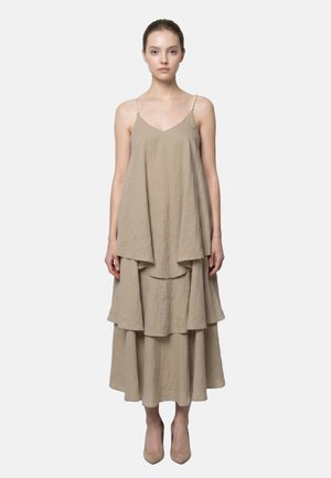 Donna che indossa un vestito senza maniche beige a strati con spalline sottili e tacchi alti beige, in piedi davanti a uno sfondo semplice.