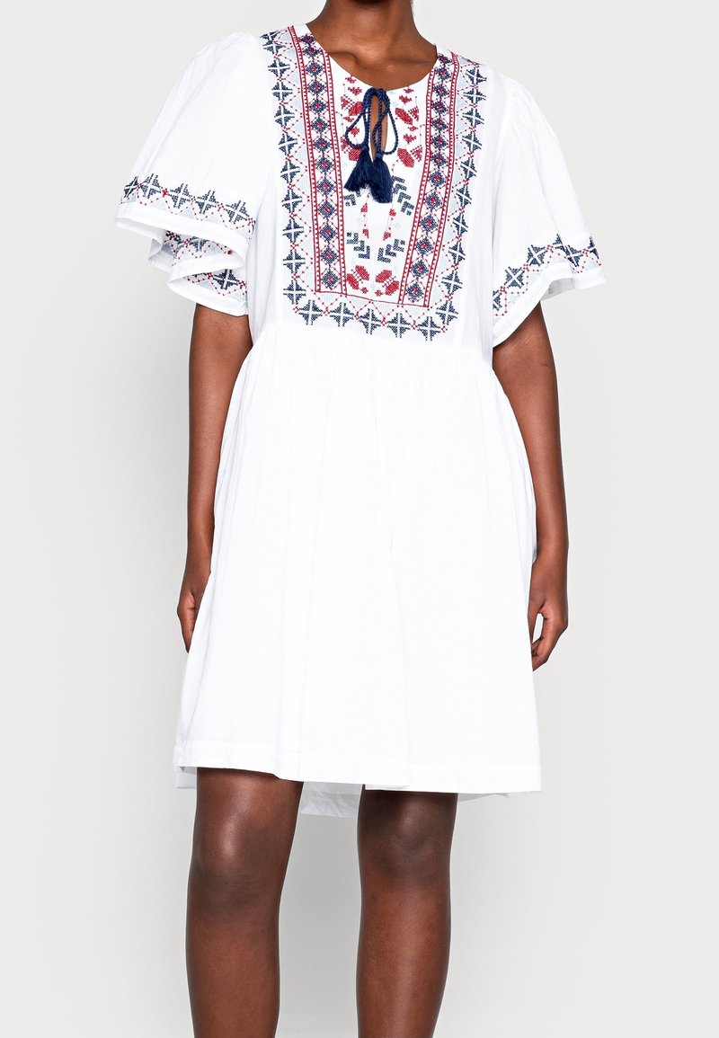 Femme portant une robe blanche jusqu'aux genoux ornée de motifs géométriques brodés en rouge et bleu avec des manches larges à volants.