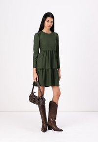 Zelena textured obleka z dolgimi rokavi in več nivojsko krilo. Ujemana s svetlo rjavimi usnjenimi škornji do kolen in majhno rjavo torbico.